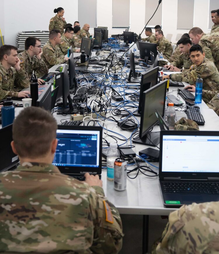 Tens of American Army soldiers sit around a table, in front of their computers