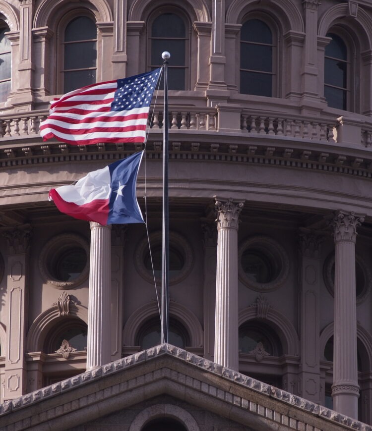 The American and Texas flags fly in front of the ornate dome and upper façade of the Texas State Capitol building in Austin, Texas.